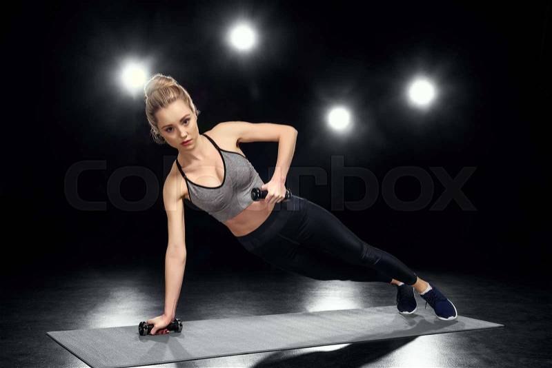 Attractive sportswoman in side plank position with dumbbells on black, stock photo