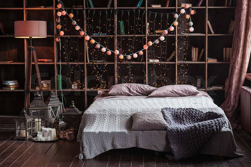 Coziness, comfort, interior and holidays concept - cozy bedroom with bed and garland lights at home. A rack with books behind the bed. Candles, a lamp and a lamp stand near the bed. Plaid hand-knitted, stock photo