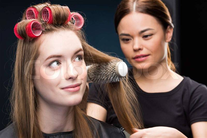 Young female hairdresser combing hair of beautiful woman in curlers, stock photo