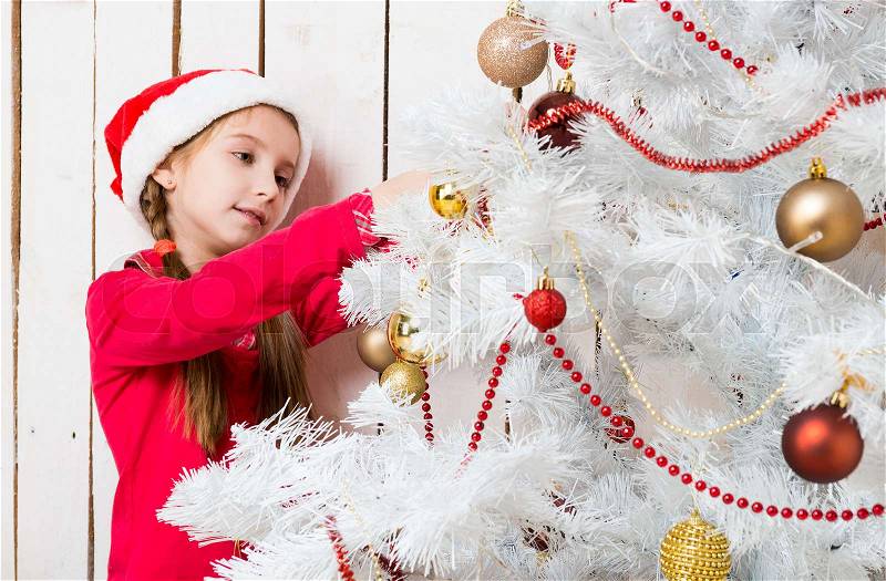 Little cute girl in red santa hat decorating new year tree, stock photo