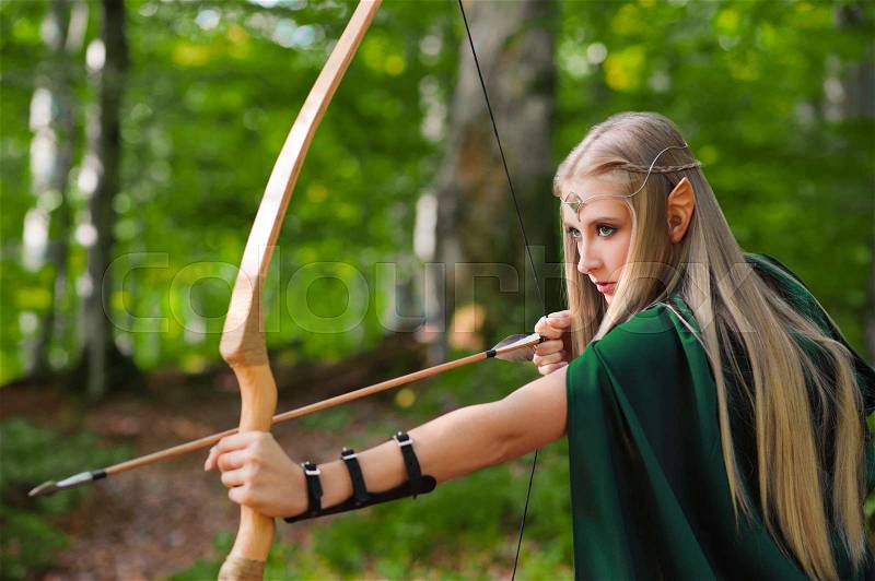 Beautiful female elf archer wearing green cape walking through the woods hunting with a bow and arrow ready to shoot danger protection defense fearless courage brave warrior fairytale fantasy costume, stock photo