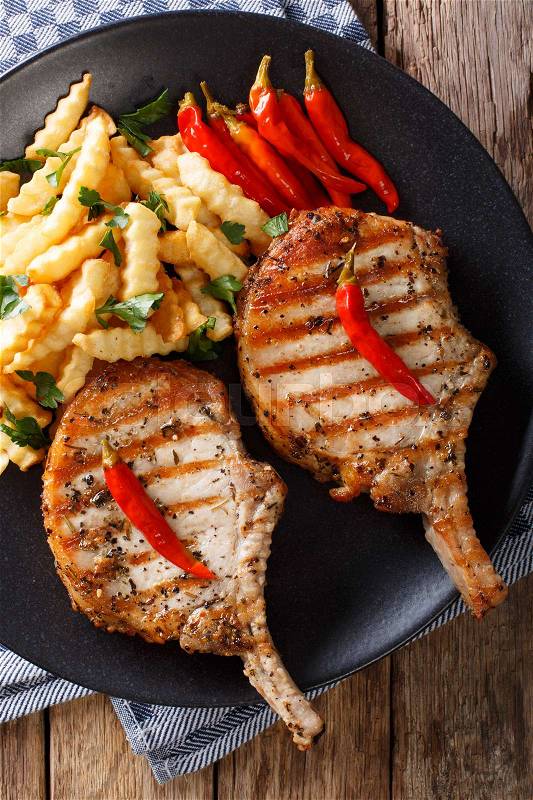 Grilled pork steak with bone, chili pepper and fries close-up on plate. Vertical view from above , stock photo