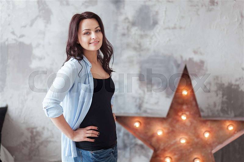 Sensual woman with dark hair in blue shirt and jeans standing near a star with light bulbs. Loft style interior, stock photo