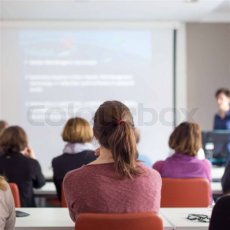 Female speaker giving presentation in lecture hall at university workshop. Rear view of unrecognized participants listening to lecture and making notes. Scientific conference event, stock photo
