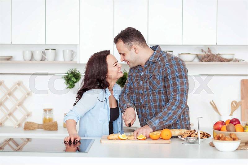 Couple making fresh organic juice in kitchen together. A young man slices a baguette. A woman standing near her boyfriend. Whiite kitchen with daylight. Healthy lifestyle concept, stock photo