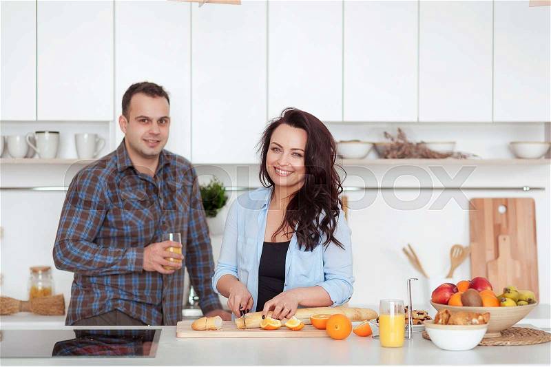 Couple making fresh organic juice in kitchen together. A young woman in a blue shirt slices a baguette. A man is hugging his girlfriend in a white kitchen. Romantic concept, stock photo