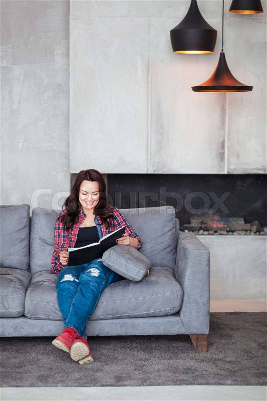Young Woman Relaxing at Home. Beautiful girl in a casual style sits on the sofa and reads a book at loft style home, stock photo