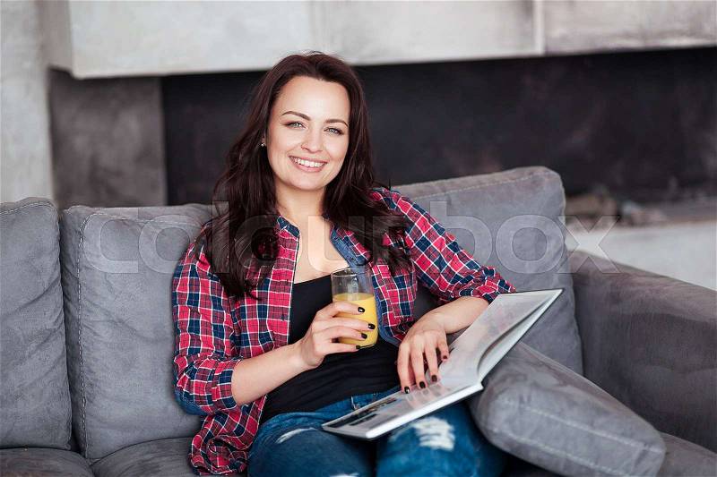Young Woman Relaxing at Home with a Glass of Orange Juice. Beautiful girl in a casual style sits on the sofa and reads a book at loft style home, stock photo