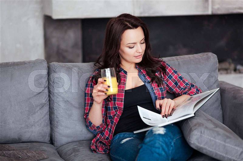 Young Woman Relaxing at Home with a Glass of Orange Juice. Beautiful girl in a casual style sits on the sofa and reads a book at loft style home, stock photo