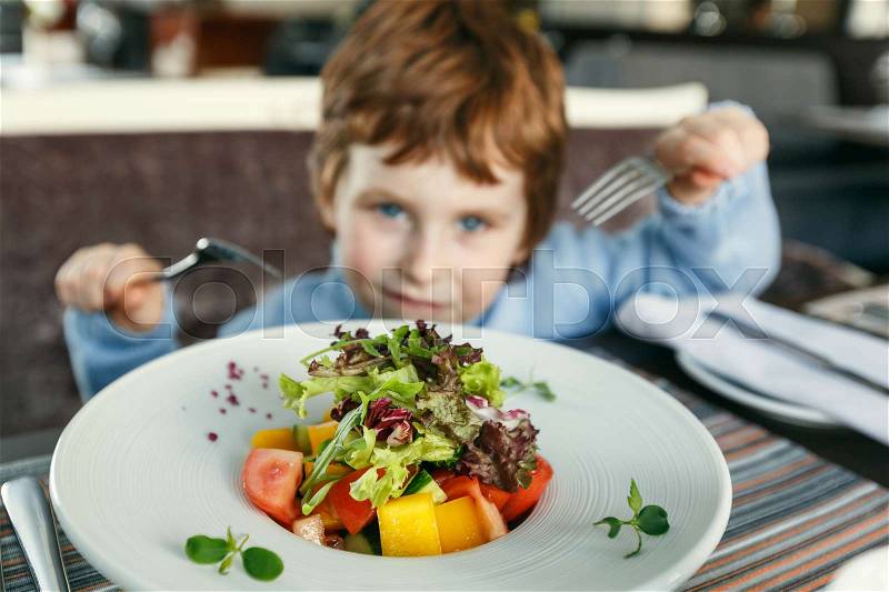 Little red haired boy with two forks eating salad in a restaurant, stock photo