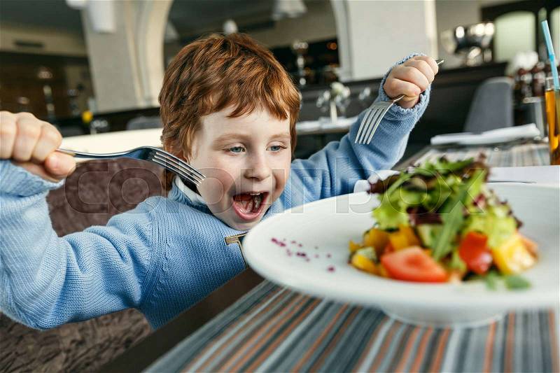 Little red haired boy with two forks eating salad in a restaurant, stock photo