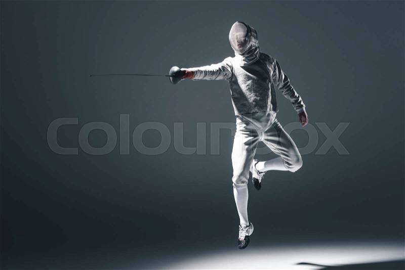 Professional fencer in fencing mask with rapier jumping on grey, stock photo