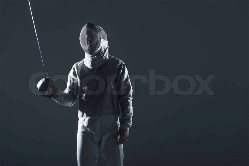 Professional fencer in fencing mask with rapier standing in position on grey, stock photo