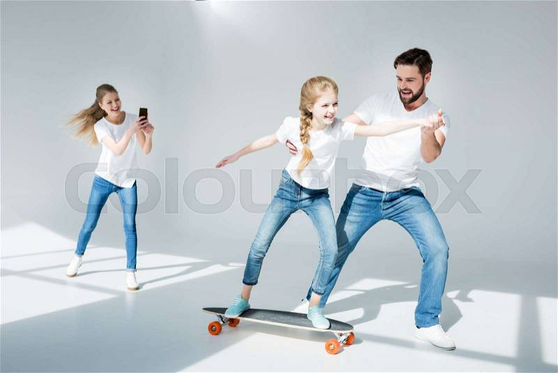 Happy woman photographing with smartphone father teaching daughter riding skateboard, stock photo