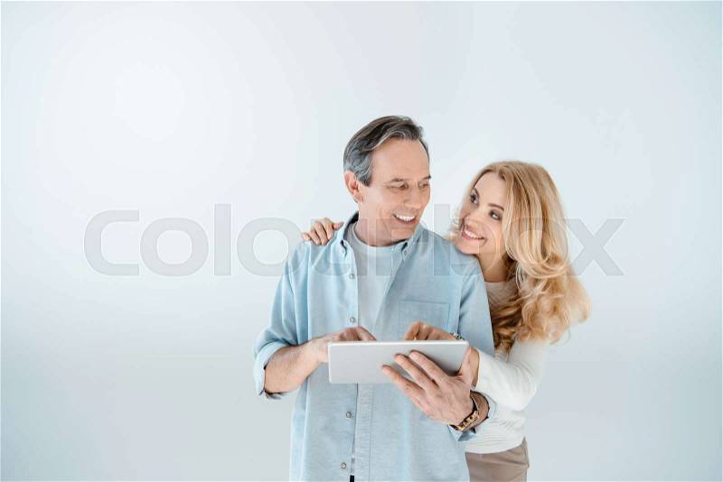 Happy middle aged couple standing embracing and using digital tablet on grey, stock photo