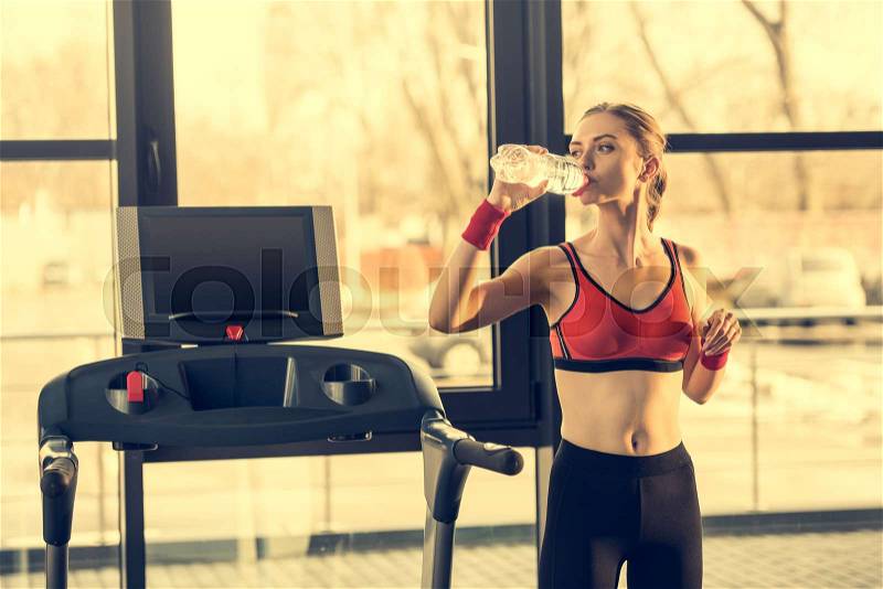 Young sporty woman drinking water from bottle while standing near trademill in gym, stock photo