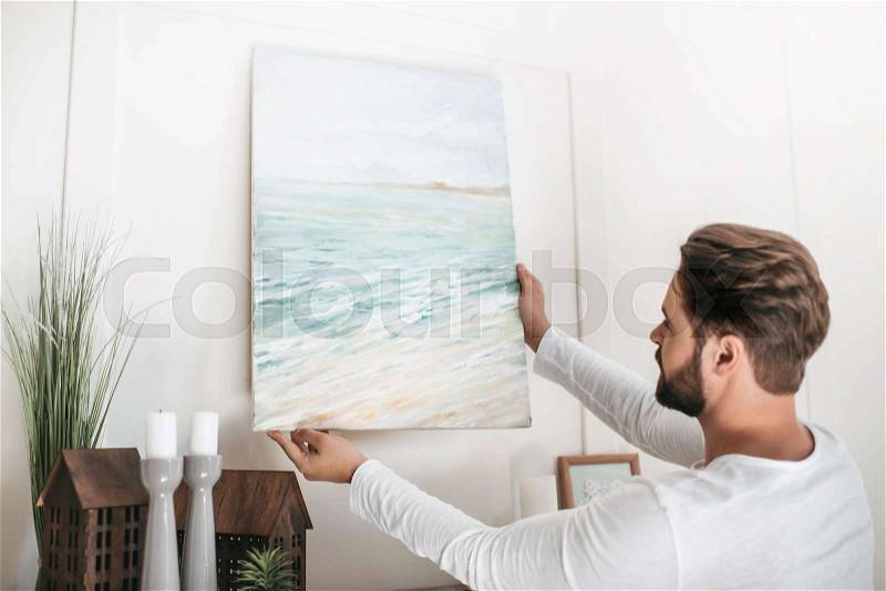 Young bearded man hanging picture on wall at home, stock photo