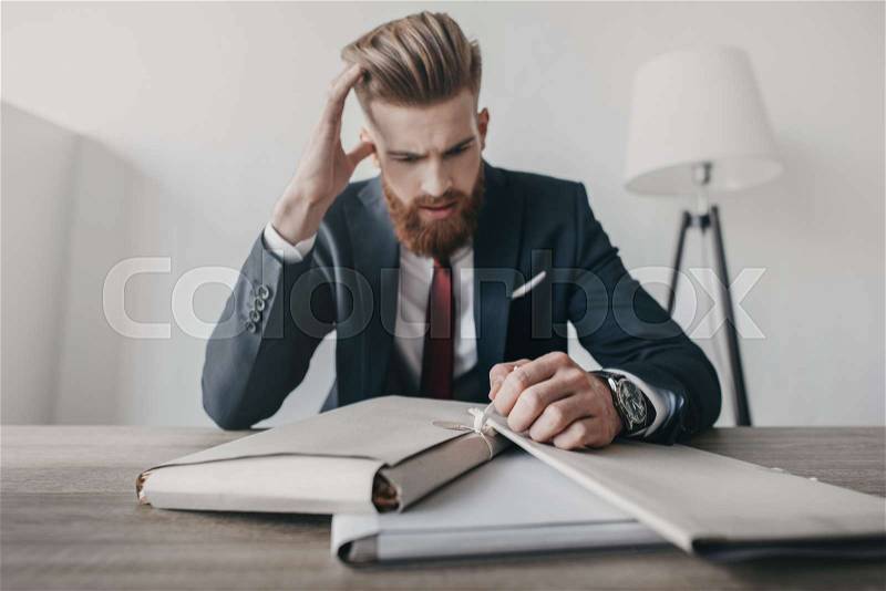 Young stressed businessman with documents and folders sitting at table in office, stock photo