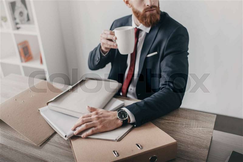 Young businessman with documents and folders drinking coffee and sitting at table in office, stock photo