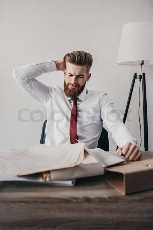Young stressed businessman with documents and folders sitting at table in office , stock photo