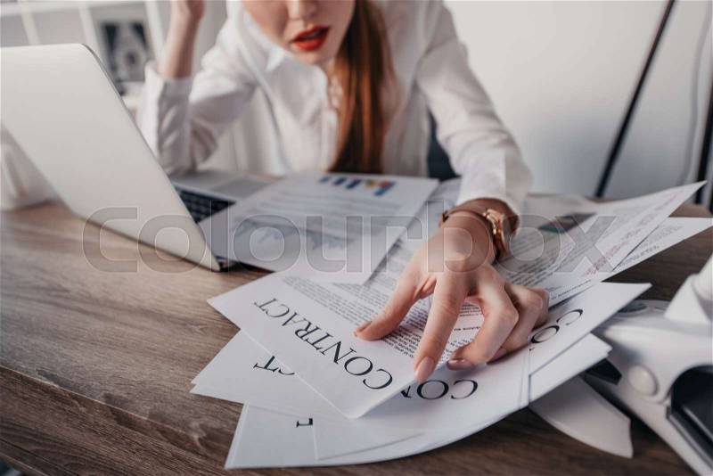 Young stressed businesswoman with laptop and contract documents sitting at table in office, stock photo