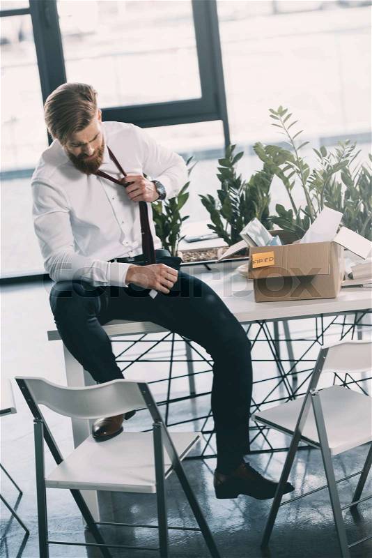 Young upset fired businessman with cardboard box sitting on table in office, stock photo