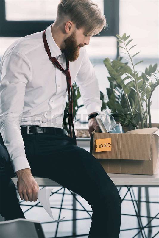 Young upset fired businessman with cardboard box in office, stock photo