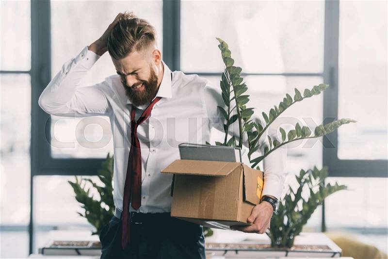 Young upset fired businessman with cardboard box in office, stock photo