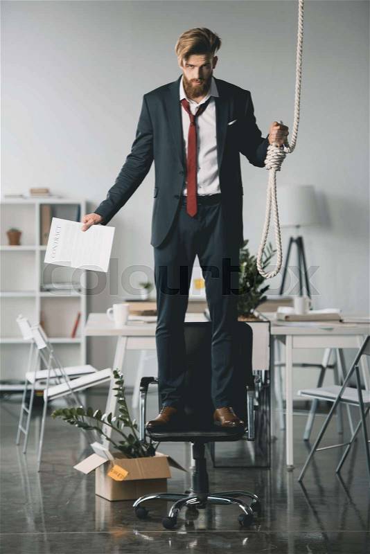 Young fired businessman standing on chair and trying to hang himself in office, stock photo