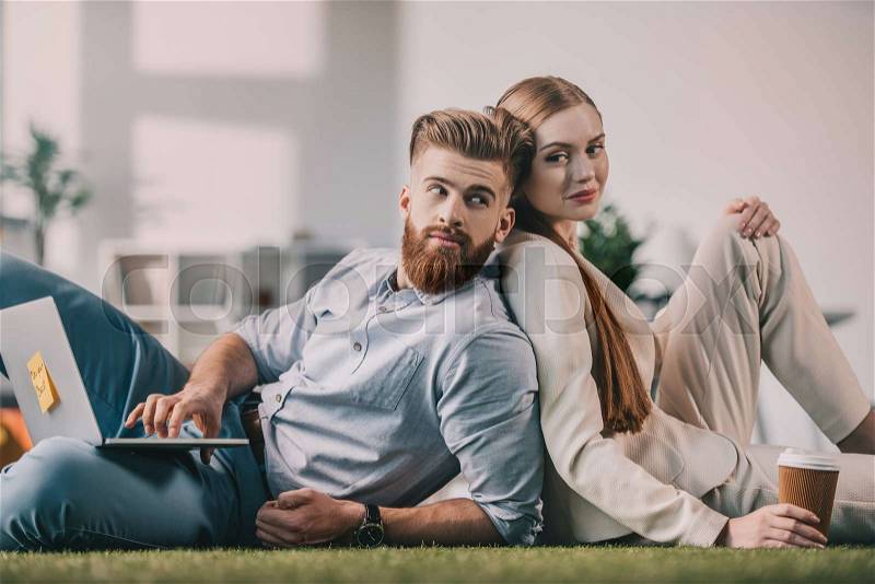 Young businessman and businesswoman sitting back to back on floor in office, stock photo