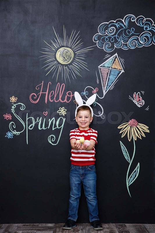 Small little kid wearing bunny ears and holding bunch of easter eggs over chalk board with colorful text background, stock photo