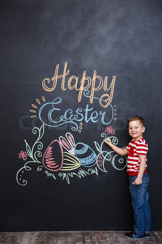 Smiling cute little boy standing and drawing easter doodles on chalk black board and looking at camera, stock photo