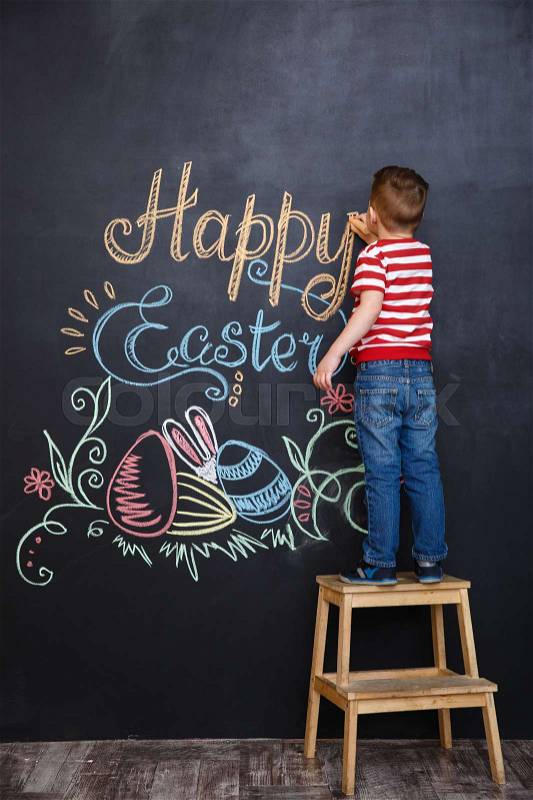 Little boy drawing easter doodles on chalk black board while standing on the ladder, stock photo