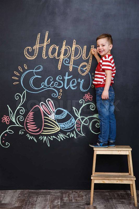Smiling cute little boy drawing easter doodles on chalk black board while standing on the ladder and looking at camera, stock photo