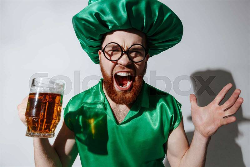 Screaming man in st.patriks costume and eyeglases which holding cup of beer in hand over gray background, stock photo