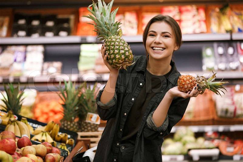 Photo of happy young lady standing in supermarket holding pineapple. Looking at camera, stock photo