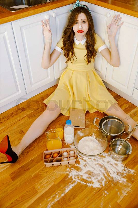 Picture of confused young pin-up woman with red lipstick sitting on floor at kitchen and cooking. Looking at camera, stock photo
