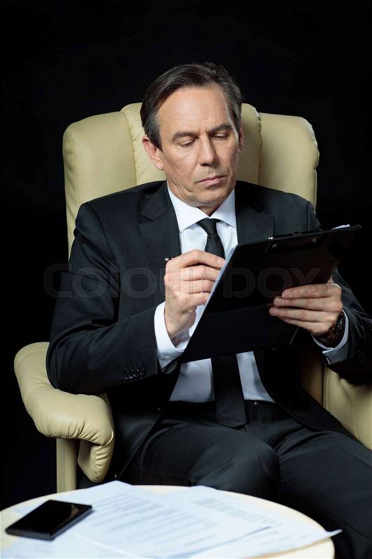 Serious mature businessman with clipboard sitting in chair and signing papers, stock photo
