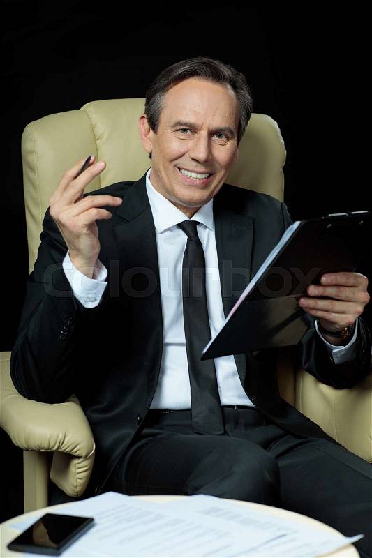 Smiling mature businessman sitting in chair and holding clipboard with papers, stock photo