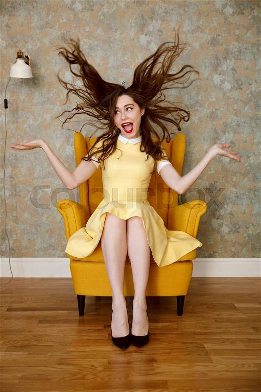 Vertical image of crazy woman in yellow dress which sitting on armchairand playing with hair while looking at camera, stock photo