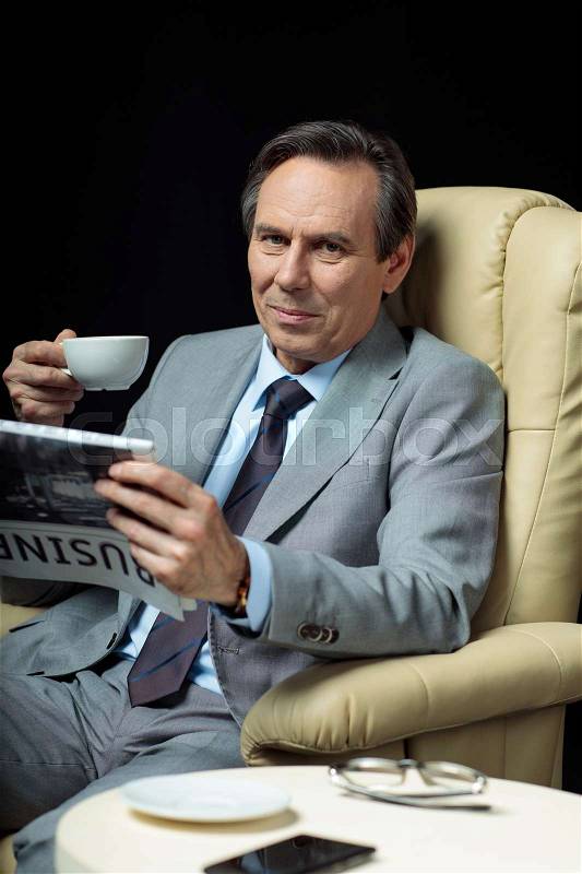 Middle aged businessman with coffee and newspaper sitting in armchair on black, stock photo