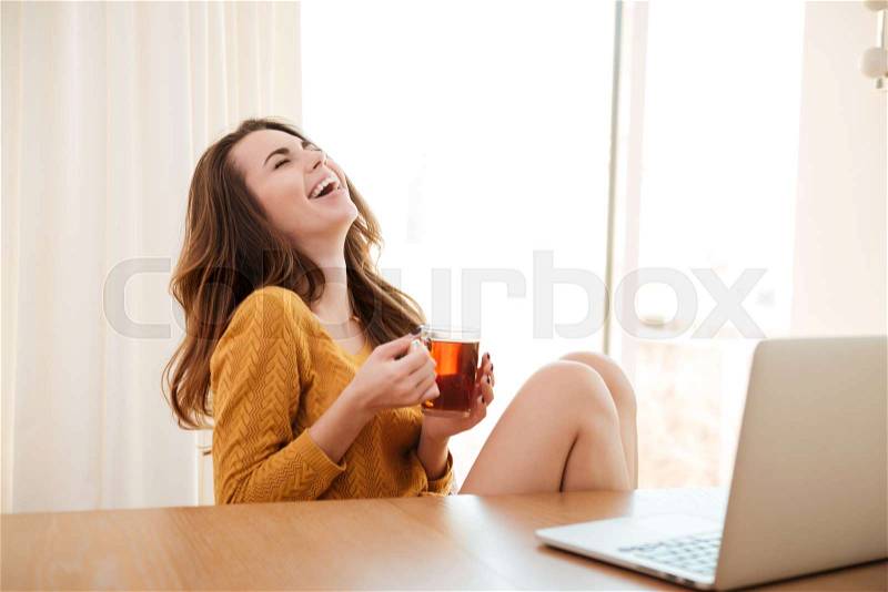 Portrait of a young cute girl in sweater holding cup of tea and laughing while sitting at the table and working at home with laptop, stock photo