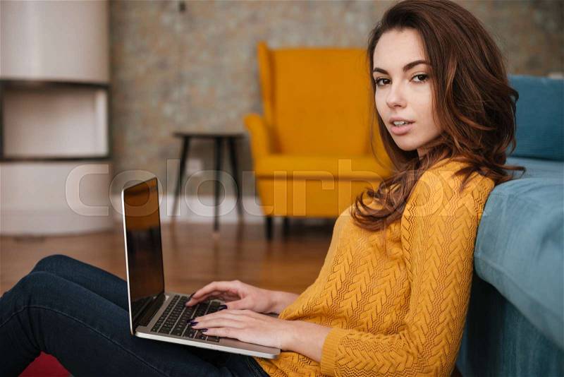 Young pretty woman sitting on floor leaning on couch and typing on laptop, stock photo