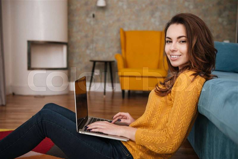 Smiling pretty woman sitting on floor leaning on couch and using laptop, stock photo