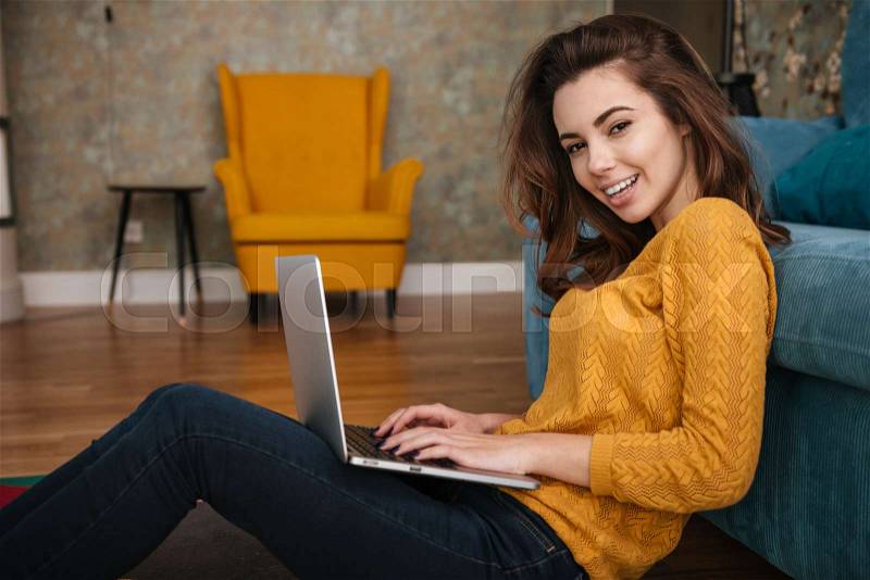 Portrait of a smiling pretty woman sitting on floor leaning on couch and using laptop, stock photo