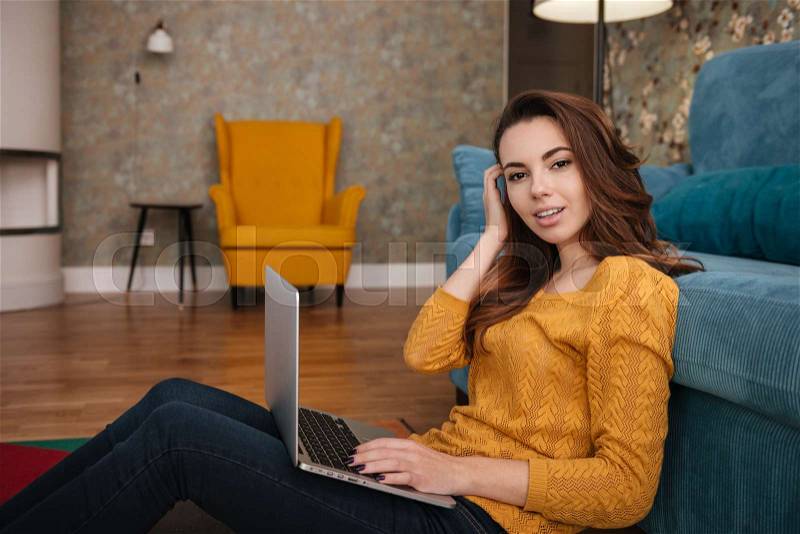 Side view of a smiling pretty woman sitting on floor leaning on couch and using laptop, stock photo
