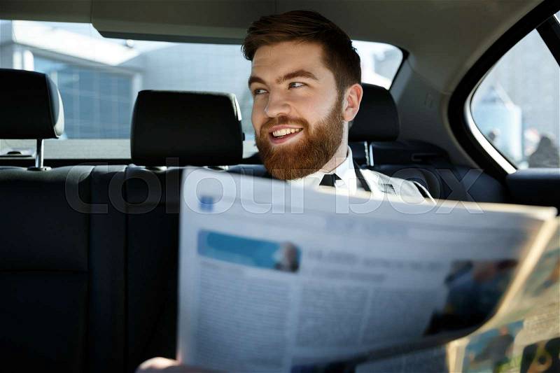 Happy bearded business man sitting on back seat of a car with newspaper and looking away, stock photo