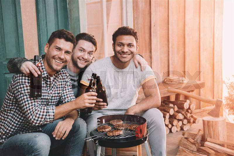 Happy young friends making barbecue and drinking beer on porch with back light, stock photo