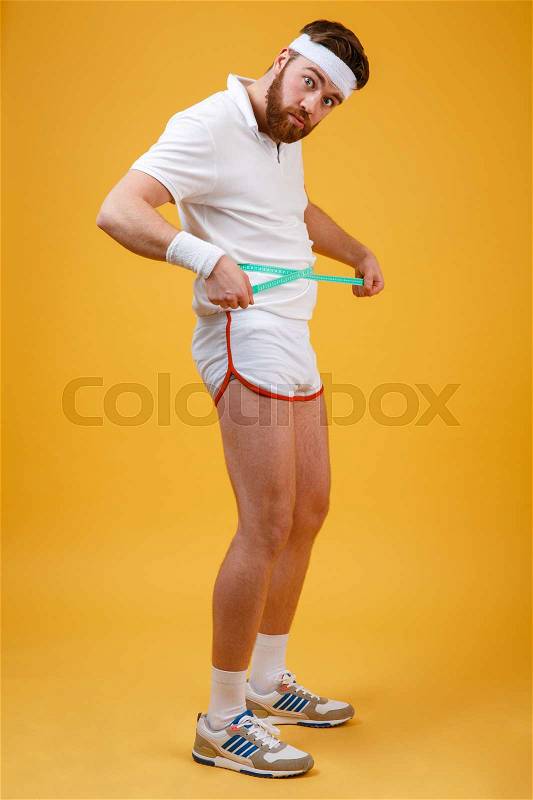 Full length portrait of a frustrated fitness man measuring his waist isolated on a orange background, stock photo