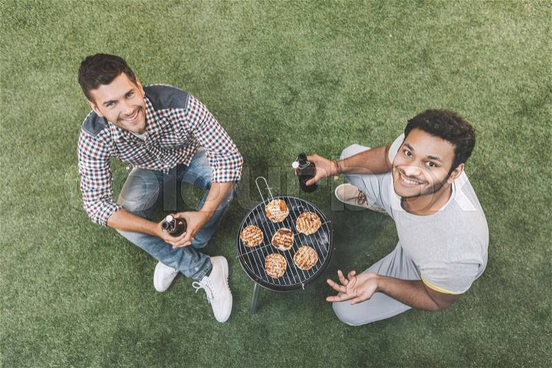 Overhead view of happy young men sitting on grass with beer bottles and grilling meat , stock photo